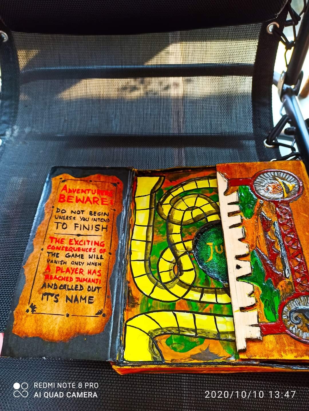 The handmade Jumanji game open — left side shows the game board with a snake path in yellow and green; right side shows the warning card "Adventurers Beware"