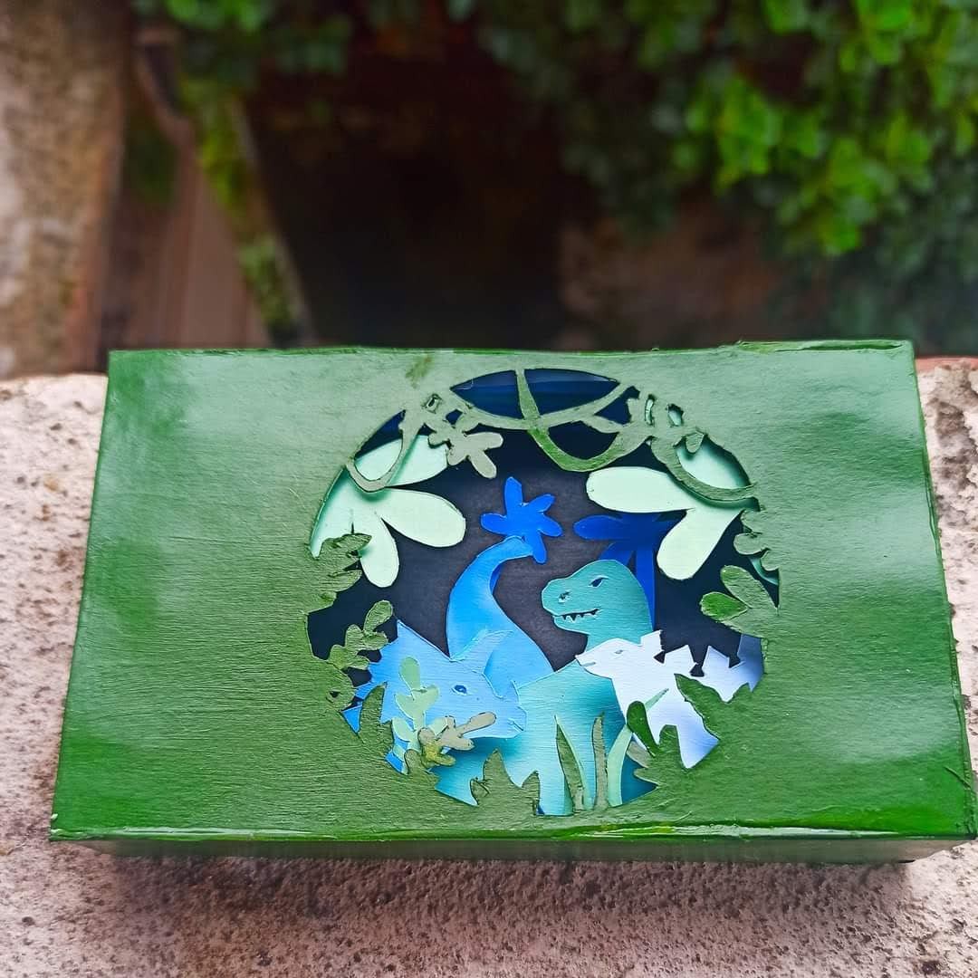 Green Amazon box with a circular portal cut in the face, revealing a layered dinosaur scene inside — T-rex and brachiosaurus in blue and teal