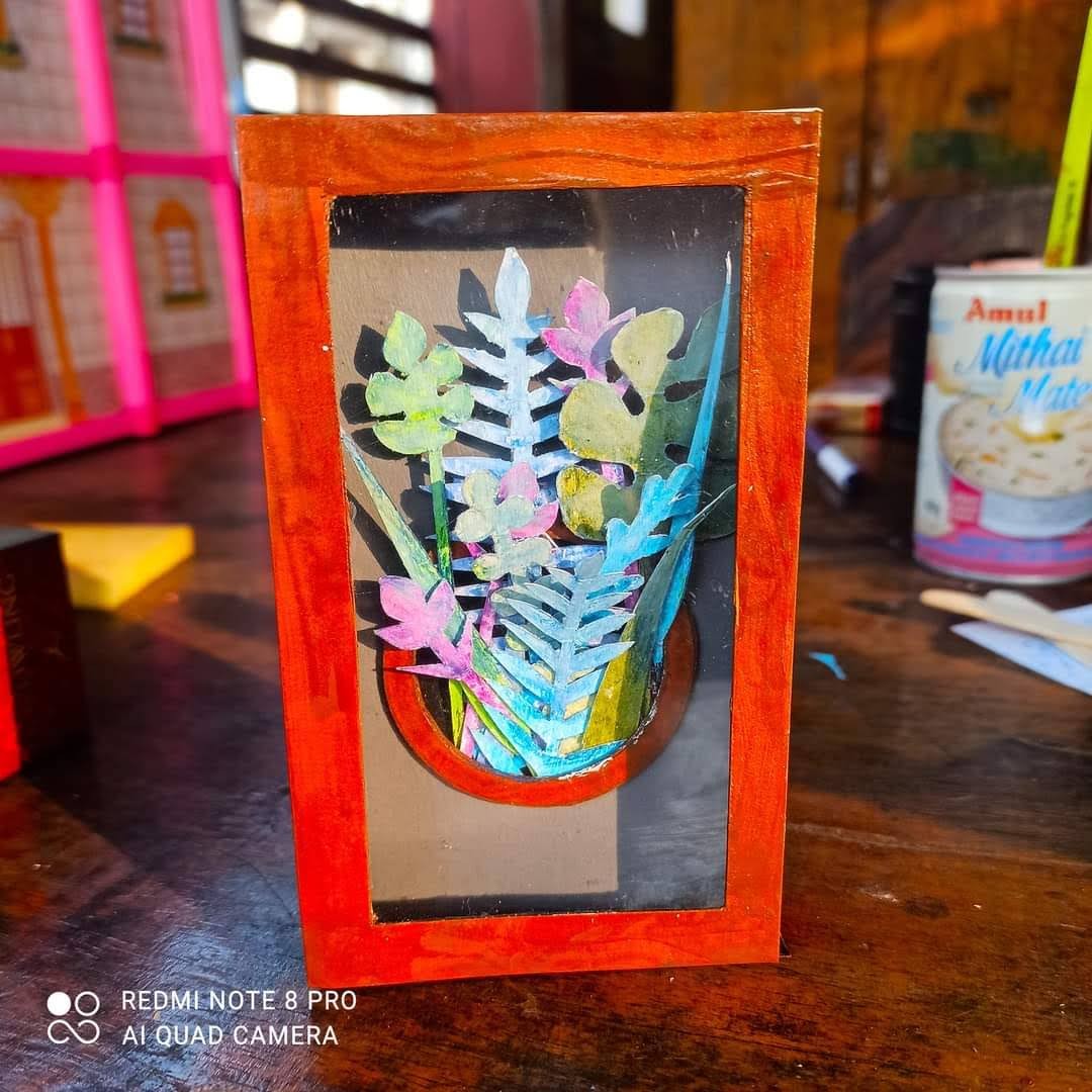Red-framed shadow box containing paper flowers and plants in a pot — pink, blue and green painted paper blooms