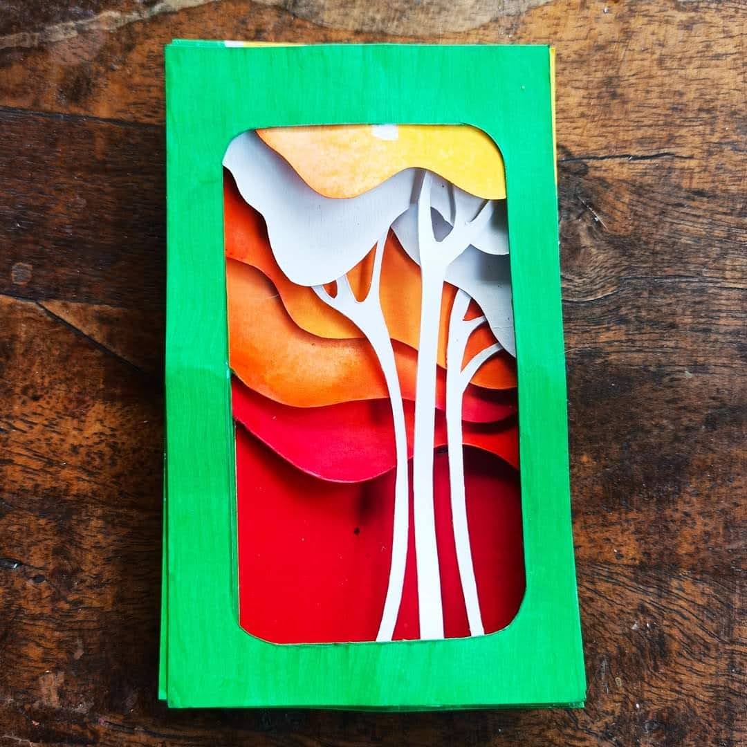 Multi-layer paper-cut forest scene in a green Amazon box frame — warm orange, red and white tree shapes receding into depth
