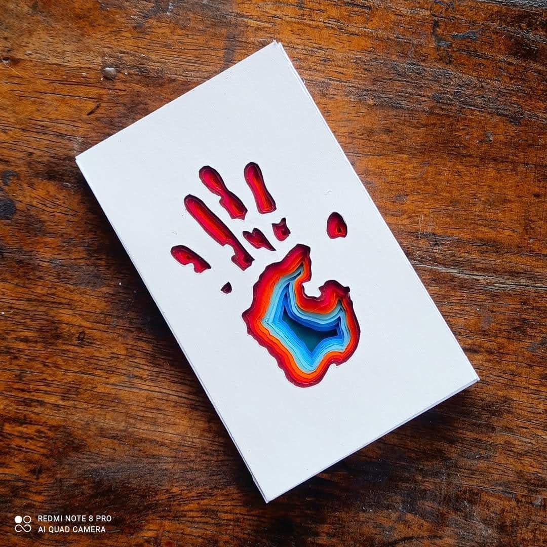 White cardboard with a handprint cut-out revealing rainbow-coloured layers below — red, orange, blue gradient at the palm centre
