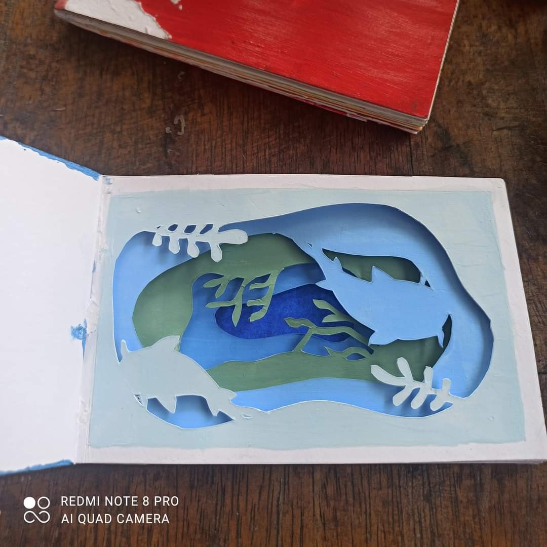 Layered paper/cardboard ocean scene — light blue, teal and green layers with dolphin silhouettes, seaweed and coral shapes