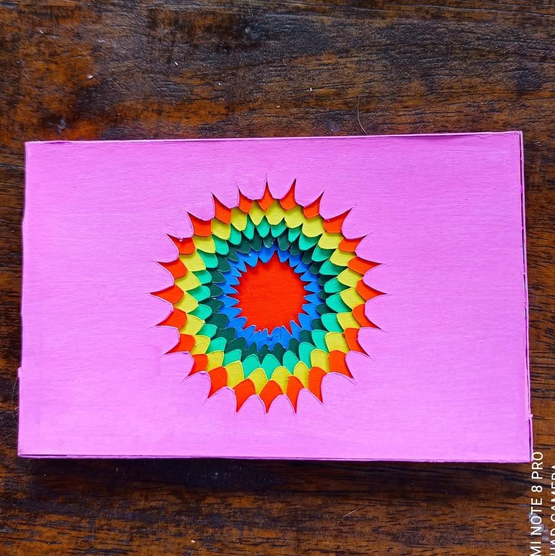 Pink cardboard with a rainbow sunburst/mandala die-cut from the centre — concentric rings of red, orange, yellow, green, teal, blue petals