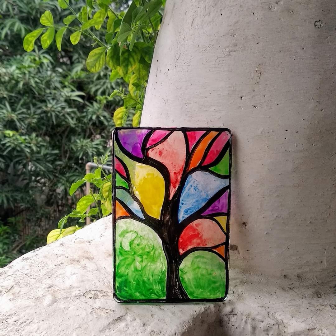 Vertical rectangular resin slab leaning against concrete — stained-glass style tree with multi-coloured canopy panels (purple, pink, orange, yellow, green, blue), black wire outlines